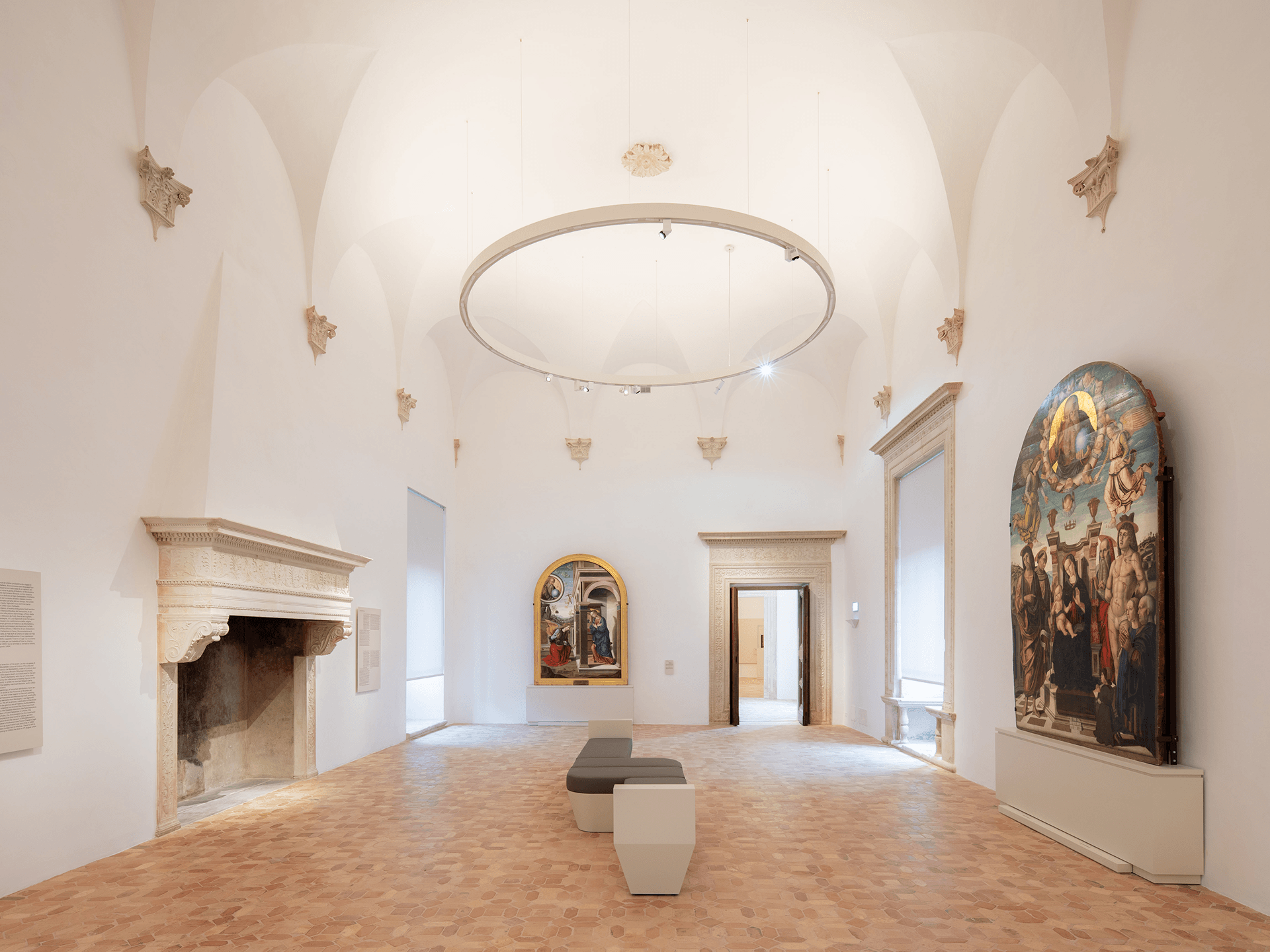 Grande ambiente museale con soffitti alti a volta, camino monumentale in pietra, sedute centrali e grandi dipinti sacri collocati lungo le pareti.