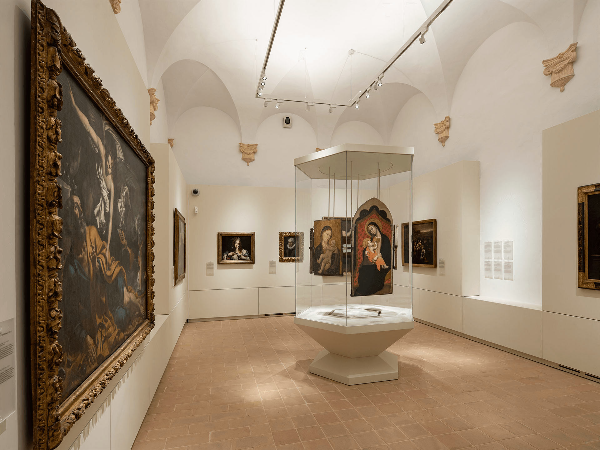 Interno di una sala museale con dipinti antichi alle pareti e una teca centrale contenente tavole sacre raffiguranti la Madonna con Bambino.