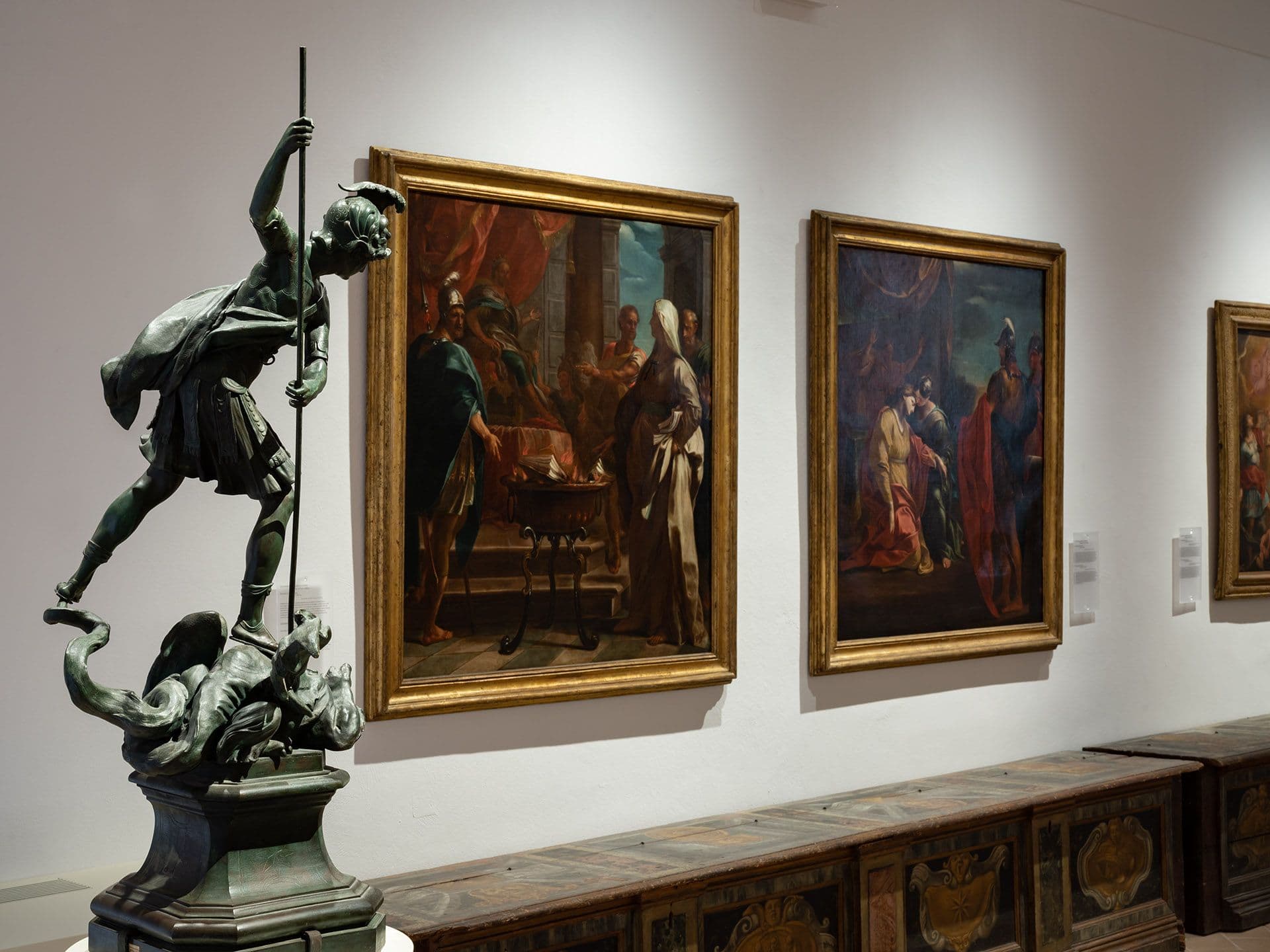 Sala museale con una scultura bronzea raffigurante un guerriero armato di lancia accanto a grandi dipinti storici incorniciati esposti lungo una parete bianca.