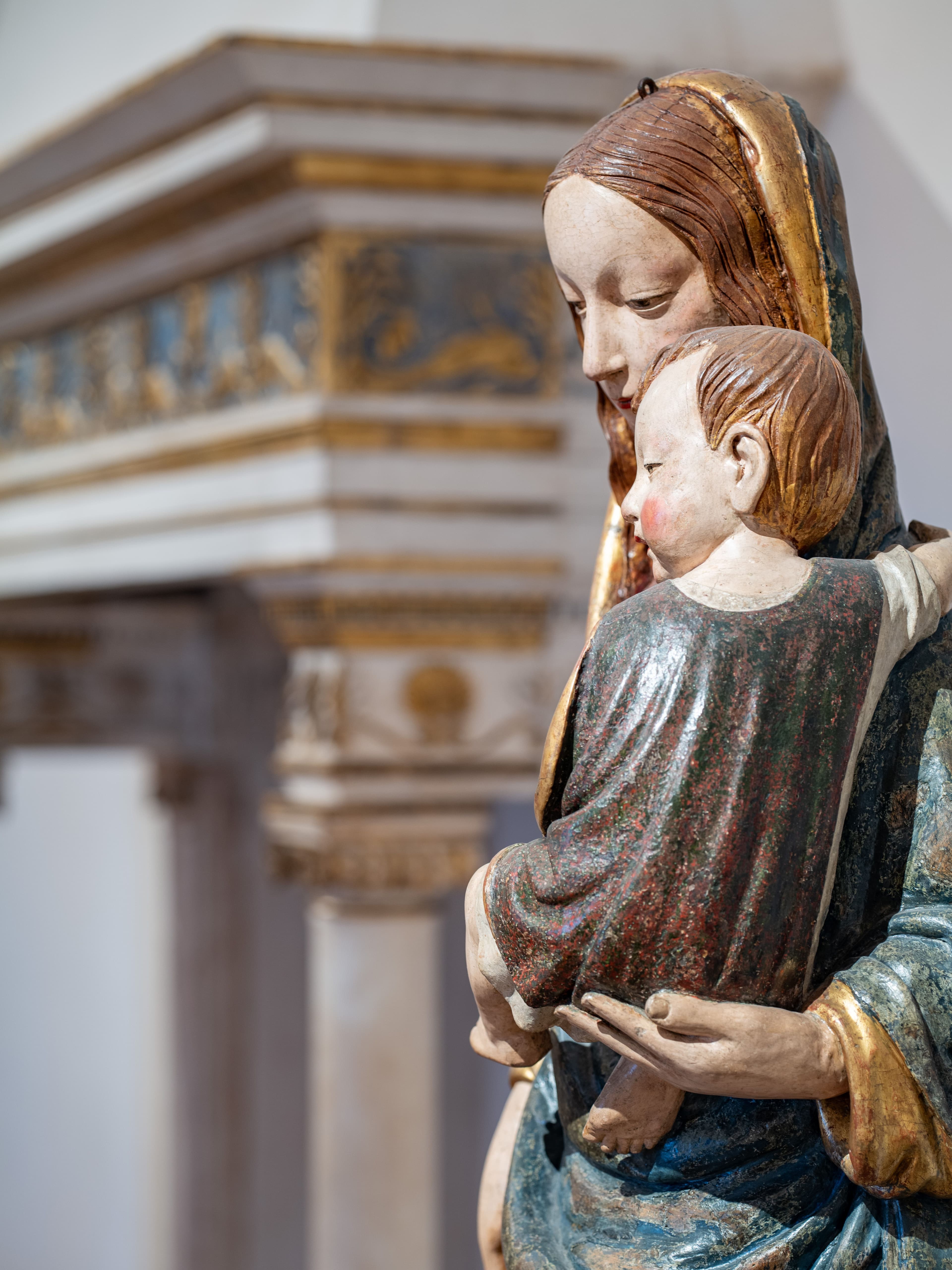 Dettaglio di una scultura lignea policroma raffigurante la Madonna con il Bambino in braccio, con sfondo sfocato di elementi architettonici decorati del museo.