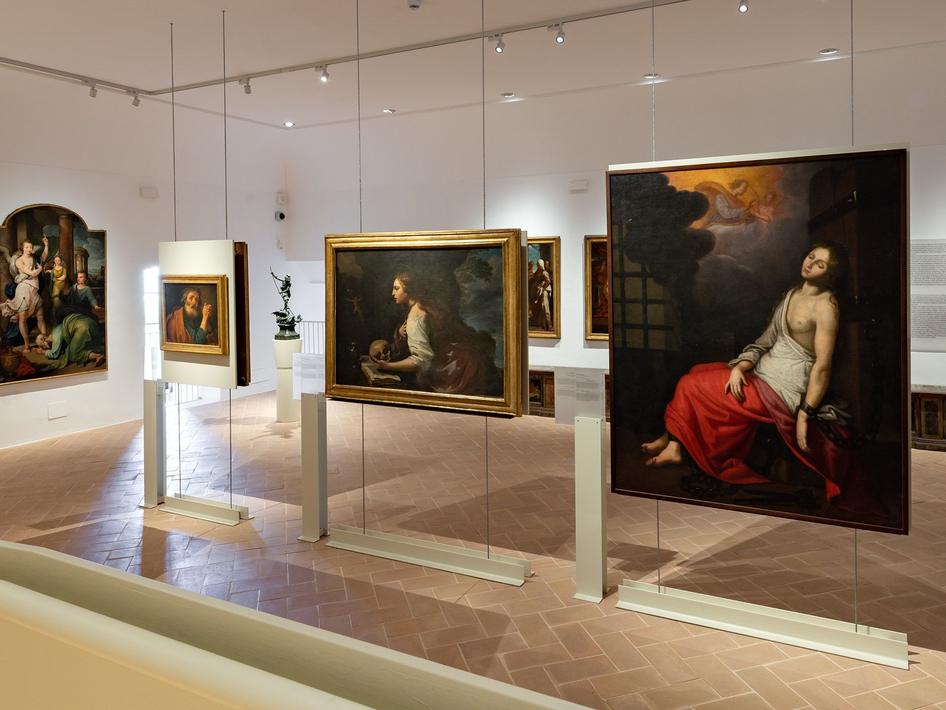 Interno di una sala museale luminosa con grandi dipinti antichi sospesi su strutture espositive e opere sacre e ritratti disposti lungo le pareti.